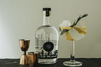 Exeter Gin bottle next to 2 rose gold jiggers and a gin goblet with a lemon twist and rosemary