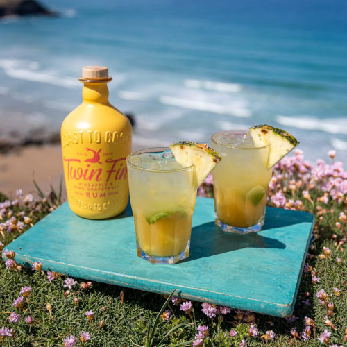 Twin Fin Pineapple and Pink Grapefruit Rum bottle next to 2 Caipirinhas on a grassy mound with a beach background