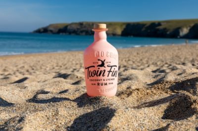 Twin Fin coconut and Lychee Rum bottle on the sand with the sea in the background