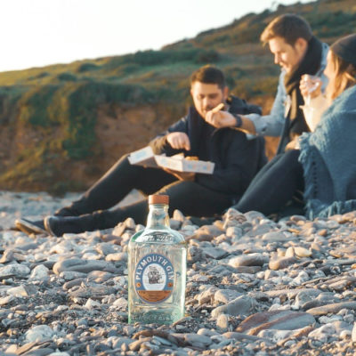 Plymouth Gin bottle on a pebbled beach with people eating fish and chips in the background